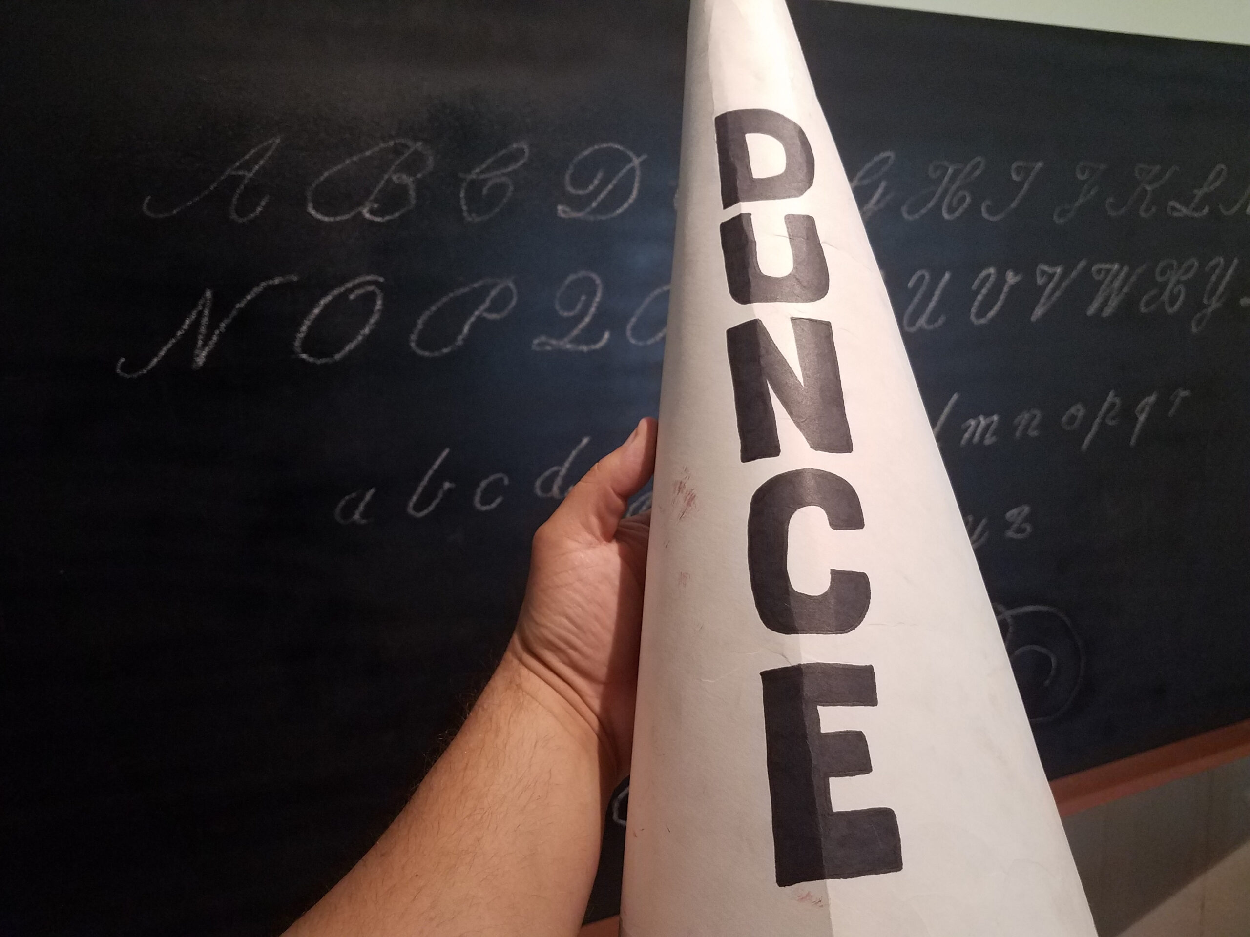 hand holding dunce cap hat in front of chalkboard or blackboard