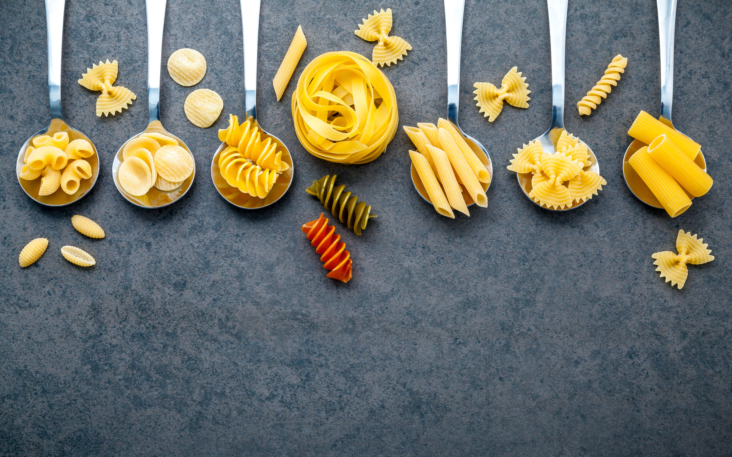 Italian foods concept and menu design. Various kind of Pasta Farfalle, Pasta A Riso, Orecchiette Pugliesi, Gnocco Sardo and Farfalle in metal spoons setup on stone background with flat lay.