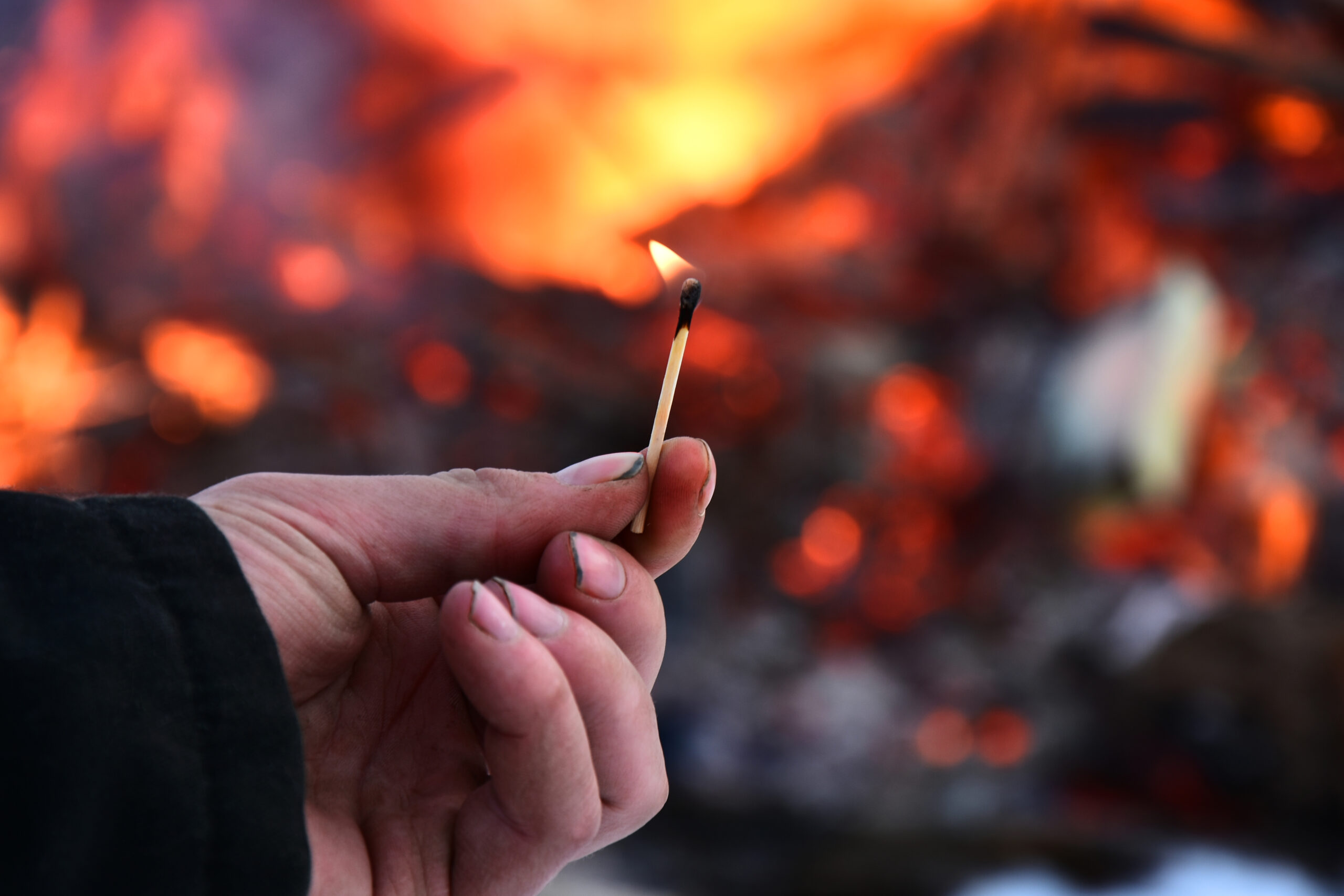 An image of a man's hand holding an open flame with a large fire in the background.